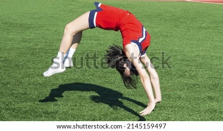 High school cheerleader warming up doing back tumbles and flips with her shadow on a green turf field before her performance.
