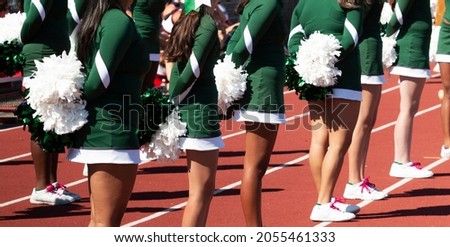 High school cheerleaders standing on a track holding their white pom poms behind their backs during a football game.