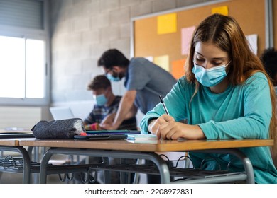 High School Female Student Wearing Face Mask In Class.doing Homework. Teacher Helping Male Teen Student In Background.