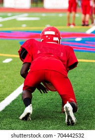 A High School Football Player At Practice In A Three Point Stance On A Green Turf Field In All Red Clothing, 