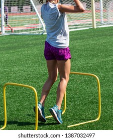 A High School Gilrl Is Taking Off To Jump Over A Two Foot High Yellow Training Hurdle During Strength And Agility Practice On A Green Turf Field.