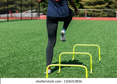 A high school girl is performing speed and agility drills over hurdles with no shoes on, only socks on a green turf field wearing black spandex, viewed from behind. - Powered by Shutterstock