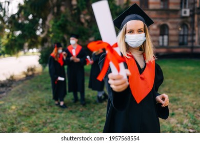 High School Graduation. Beautiful Female Graduate In A Medical Protective Mask On Her Face And A Hat On Her Head, Outdoors, Shows A Diploma With A Red Ribbon. Distance Learning, Coronavirus.