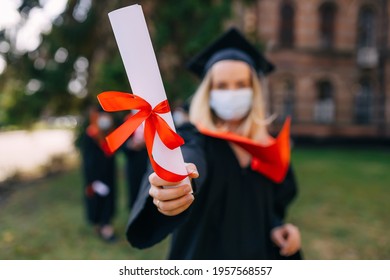 High School Graduation. Beautiful Female Graduate In A Medical Protective Mask On Her Face And A Hat On Her Head, Outdoors, Shows A Diploma With A Red Ribbon. Distance Learning, Coronavirus.