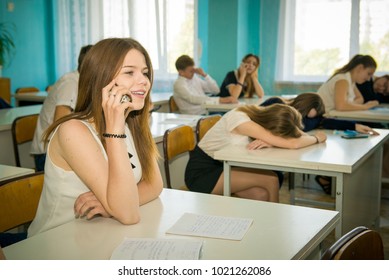 High School Student At A Desk With A Mobile Phone. Girl Communicates On The Phone At A Lesson At School. Behavior Of Modern Youth In School. Blurred Background, No Model Release