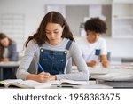 High school student taking notes from book for her study. Young woman sitting at desk and finding information in college library. Focused girl studying in classroom completing assignment.