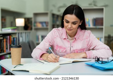 High School Student Taking Notes From Book For Her Study. Young Woman Sitting At Desk And Finding Information In College Library. Focused Girl Studying In Classroom Completing Assignment 