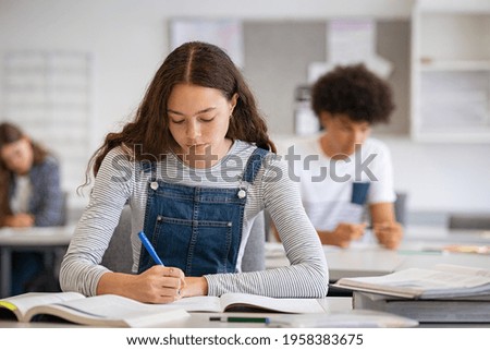 High school student taking notes from book for her study. Young woman sitting at desk and finding information in college library. Focused girl studying in classroom completing assignment.