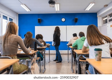 High school students are taking notes in a classroom while their teacher explains the lesson standing next to a whiteboard