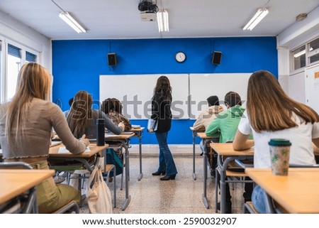 High school students are taking notes in a classroom while their teacher explains the lesson standing next to a whiteboard