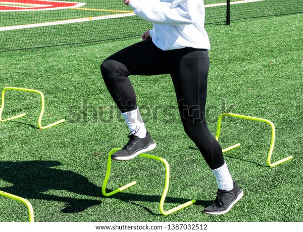 A high school teenage girl is doing a running\
drill stepping over yellow mini hurdles during track and field\
practice on a green turf\
field.