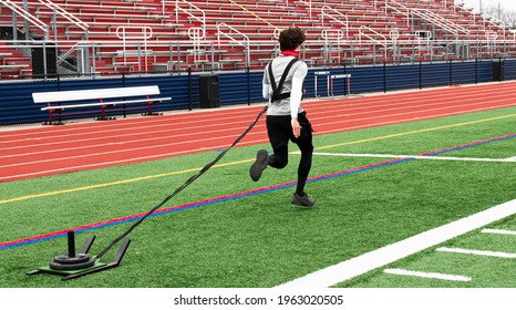 A high school track runner is pulling a sled with weights on it across a green turf field during strngth and agility practice.