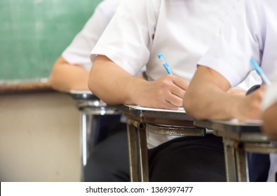 High School Or University Student Holding Pencil Writing On Paper Answer Sheet.sitting On Lecture Chair Taking Final Exam Attending In Examination Room Or Classroom.student In Uniform 