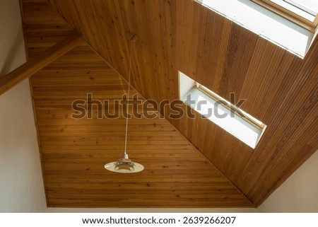 High sloping ceiling, completely covered with wooden panels. Two dormer windows are built into the ceiling, through which bright daylight penetrates. A single retro-style lamp hangs from the ceiling.
