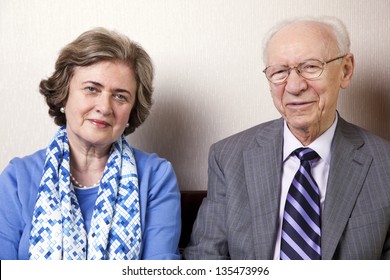 A High Society Senior Couple (he's In His 80's, She's In Her Late 60's) Sitting On A Sofa Looking At The Camera. Medium Close Up.