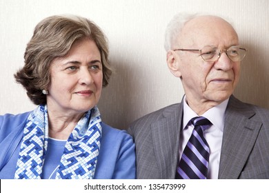 A High Society Senior Couple (he's In His 80's, She's In Her Late 60's) Sitting On A Sofa Looking Away To The Right Side Of The Frame.