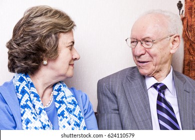 A High Society Senior Couple (he's In His 80's, She's In Her Late 60's) Sitting On A Sofa, Having A Loving Moment Together.