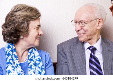 A High Society Senior Couple (he's In His 80's, She's In Her Late 60's) Sitting On A Sofa, Having A Loving Moment Together.