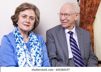 A High Society Senior Couple (he's In His 80's, She's In Her Late 60's) Sitting On A Sofa, Having A Loving Moment Together.