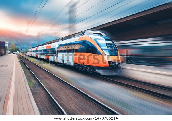 High speed orange train in motion on the\
railway station at sunset. Modern intercity passenger train with\
motion blur effect on the railway platform. Industrial. Railroad in\
Europe. Transport
