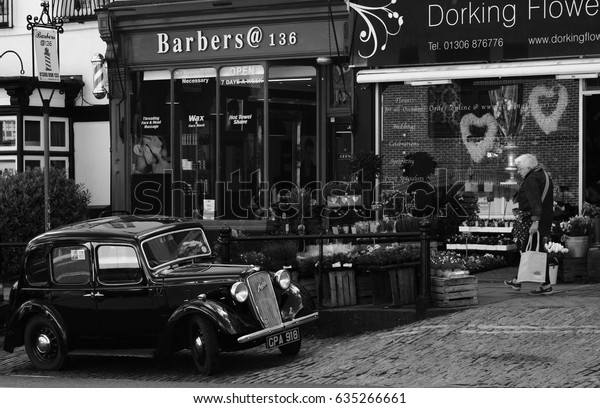 High Street, Dorking, Surrey, United\
Kingdom, 25 April, 2017: An Austin Ruby Car Parked Outside a Flower\
and Barber Shop with an Elderly Person Walking\
By.