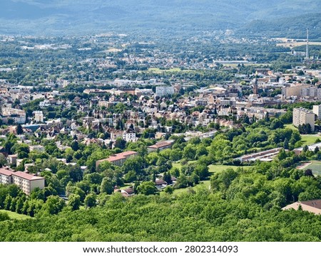 High vantage panoramic view of small European town surrounded by dense green trees and rolling hills, showing residential buildings, red tile roofs, church spire, parks, winding roads, and mountain