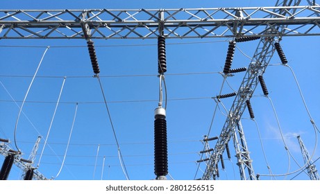 High voltage electrical substation featuring brown porcelain insulators suspended from steel lattice structures against a clear blue sky. Essential power grid distribution and industrial energy infrastructure concept.
