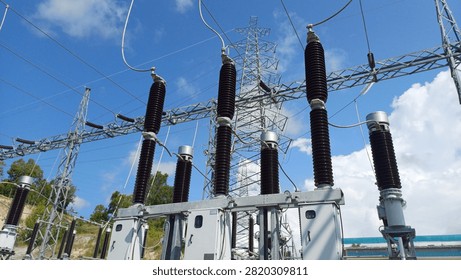 High voltage electrical substation featuring prominent current transformers with brown porcelain insulators, connected to a steel lattice transmission tower under a bright blue sky. Industrial power distribution infrastructure and electrical grid utility concept.
