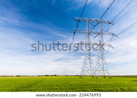High voltage lines and power pylons in a flat and green agricultural landscape on a sunny day with cirrus clouds in the blue sky.