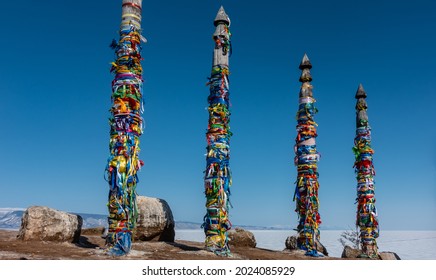 High wooden ritual pillars are tied with colorful ribbons. Bright colors against the background of a clear blue sky and white ice of a frozen lake. Picturesque boulders on the ground. Baikal