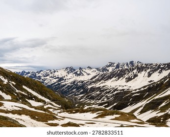 A high-altitude Alpine landscape featuring dramatic snow-covered peaks, a deep rocky valley with a winding mountain road, and high-voltage power lines under an overcast sky.