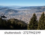 High-altitude perspective looking down over sprawling city nestled between layered mountain ridges. Evergreen trees frame the foreground. Sarajevo, Bosnia and Herzegovina