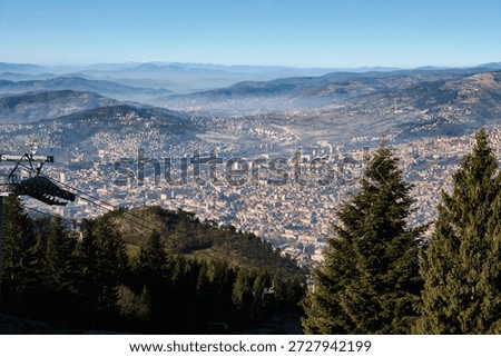 High-altitude perspective looking down over sprawling city nestled between layered mountain ridges. Evergreen trees frame the foreground. Sarajevo, Bosnia and Herzegovina