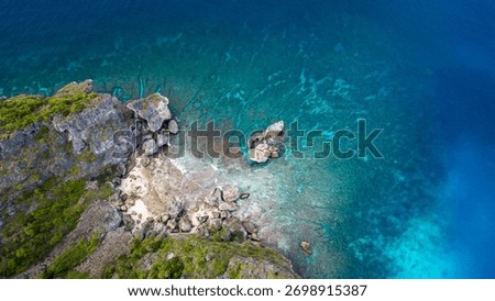 A high-angle aerial view capturing a rugged, lush green cliff face meeting a small, rocky white beach. The surrounding ocean showcases incredible clarity, with vibrant turquoise and deep blue colors