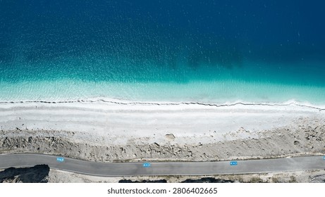 High-angle aerial view looking down at a section of the coastal road near Lake Salda in Burdur, Turkey, with blue bicycle lane symbols. A single white car is seen, emphasizing the remote nature alongside the white mineral beach and turquoise lake.