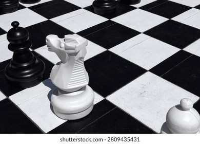 A high-angle close-up captures oversized chess pieces placed upon a large, high-contrast black and white checkered floor surface. In this outdoor environment, the sunlight creates sharp, definitive shadows, enhancing the dimensional presence of the heavy, smooth figures. Centrally positioned, a white knight stands prominently, its stylized design characterized by ribbed neck details and a distinctively sculpted equine head, casting a notable shadow onto the adjacent dark tile. To its upper left, a large, rounded black piece, likely a bishop, commands a presence on the grid, its smooth finish contrasting with the gritty texture of the flooring tiles. Further down in the lower right corner, the top portion of another white, spherical element is partially visible, hinting at the breadth of the set. The layout suggests an immersive, life-sized version of the classic strategic board game. The perspective emphasizes the scale difference between the ground tiles and the towering tokens. Every surface exhibits wear, with minor imperfections and dust scattered across the squares, lending an authentic, tactile feel to the setting. The play of light and dark geometry dominates the composition, creating a striking visual rhythm that draws the eye across the board. The stark coloration of the playing area is reflected in the monochromatic scheme of the figures, establishing a coherent theme throughout the entire space. This scene captures a moment of calculated pause in a competitive engagement, where the physical reality of the pieces feels tangible and grounded within the public or stylized venue where they reside, inviting curiosity about the tactical situation unfolding among these giants.