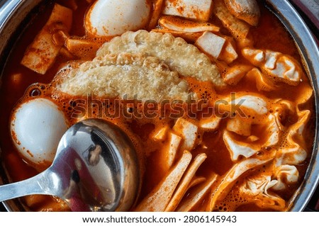 A high-angle close-up shot of appetizing Korean Tteokbokki boiling in a pot, featuring crispy fried dumplings, a boiled egg, and a silver metal ladle resting on the left side.