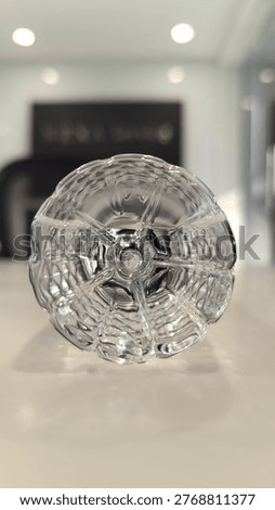 A high-angle, close-up shot of a clear, pressed glass bobeche or decorative crystal dish. The image highlights the intricate, starburst-like geometric patterns and scalloped edges, creating a sense of symmetry and elegance through light refraction on a clean, neutral background.