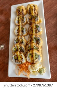 High-angle, close-up shot of crispy fried tempura sushi rolls arranged on a white rectangular plate. The rolls are drizzled with savory sauce and served with fresh shredded carrots, pickled ginger, and a dollop of green wasabi on a dark wooden table background. Professional food photography
