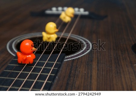 A high-angle close-up shot featuring one orange and two yellow plastic ducks sitting on the strings of a dark wooden acoustic guitar. This whimsical image symbolizes creativity, music lessons for children, and the joy of learning an instrument in a fun, artistic way. The shallow depth of field highlights the leading orange duck.