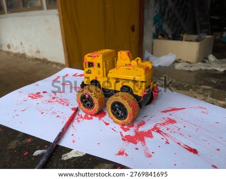 A high-angle, close-up shot featuring a yellow plastic construction toy truck heavily splattered with vibrant pink paint. The truck sits on a white sheet of paper covered in messy paint streaks and tire tracks, illustrating a creative art session or sensory play activity. A thin paintbrush rests in the foreground, suggesting an ongoing artistic process. The natural outdoor lighting highlights the glossy texture of the wet paint against the rugged tires of the toy. This image captures themes of childhood creativity, artistic exploration, and the 
