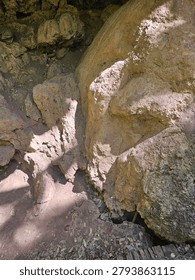 A high-angle, close-up shot looking down into a rugged stone alcove or cavern entrance. Harsh, bright sunlight streams from the upper right, illuminating the rough, porous surface of large tan boulders while casting deep shadows into the rocky crevices. The dirt floor below is scattered with dry, fallen leaves, highlighting the natural, raw texture of a wilderness cave environment.