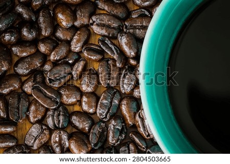 A high-angle close-up shot of roasted coffee beans scattered on a wooden surface next to a teal cup filled with black coffee, illustrating a thematic focus on coffee culture and morning rituals.