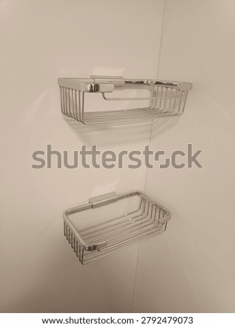A high-angle, close-up shot of a set of two modern chrome-finished wire shower baskets mounted on a clean, off-white corner wall in a contemporary bathroom, as captured in image. These sleek, rectangular stainless steel soap and shampoo holders feature a minimalist metal wire design that allows water to drain efficiently. This functional bathroom hardware represents neat home organization, luxury restroom amenities, modern interior design, and household cleanliness. Perfect stock imagery for home improvement blogs, interior design portfolios, retail product catalogs, and sanitary ware marketing materials.