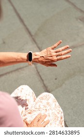A high-angle close-up view of an elderly woman's wrinkled hand and wrist wearing a modern smart fitness tracker. The electronic wearable device is shown outdoors in bright daylight.