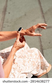 A high-angle close-up view of an elderly woman's wrinkled hands adjusting a modern smart fitness tracker on her wrist. The electronic wearable device is shown outdoors in bright daylight.