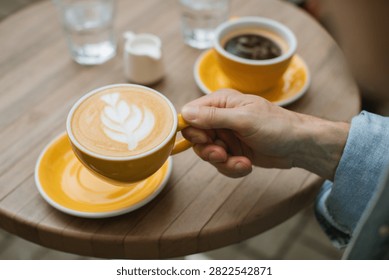 High-angle close-up view of a man's hand holding a yellow cup with rosette latte art over a saucer, with another cup of black coffee and water glass on a round wooden table.