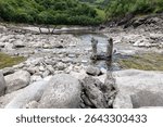 High-angle, close-up view of a rocky streambed with shallow, clear water flowing through it. The stream is narrow, and the bed is composed primarily of various sizes of grey and light-colored