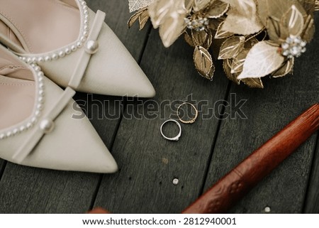 A high-angle composition featuring white bridal heels decorated with pearls, two wedding rings, a traditional golden wedding headpiece, and a wooden Malay keris on a dark wooden floor.