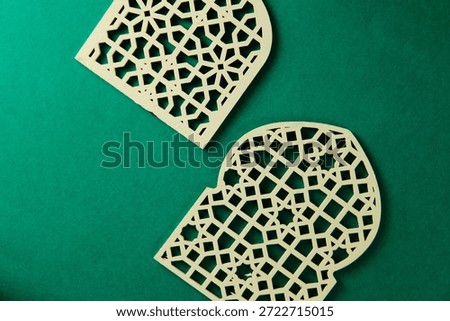 A high-angle, creative still life featuring intricate Islamic-themed decorations. The composition includes a laser-cut wooden lantern (Fanous) with internal LED lighting and two decorative wooden panels with geometric mashrabiya patterns. Set against a vibrant emerald green backdrop, the warm wood tones and glowing light create a peaceful, celebratory atmosphere. Ideal for Ramadan Kareem, Eid al-Fitr, or Islamic heritage marketing campaigns and social media content.